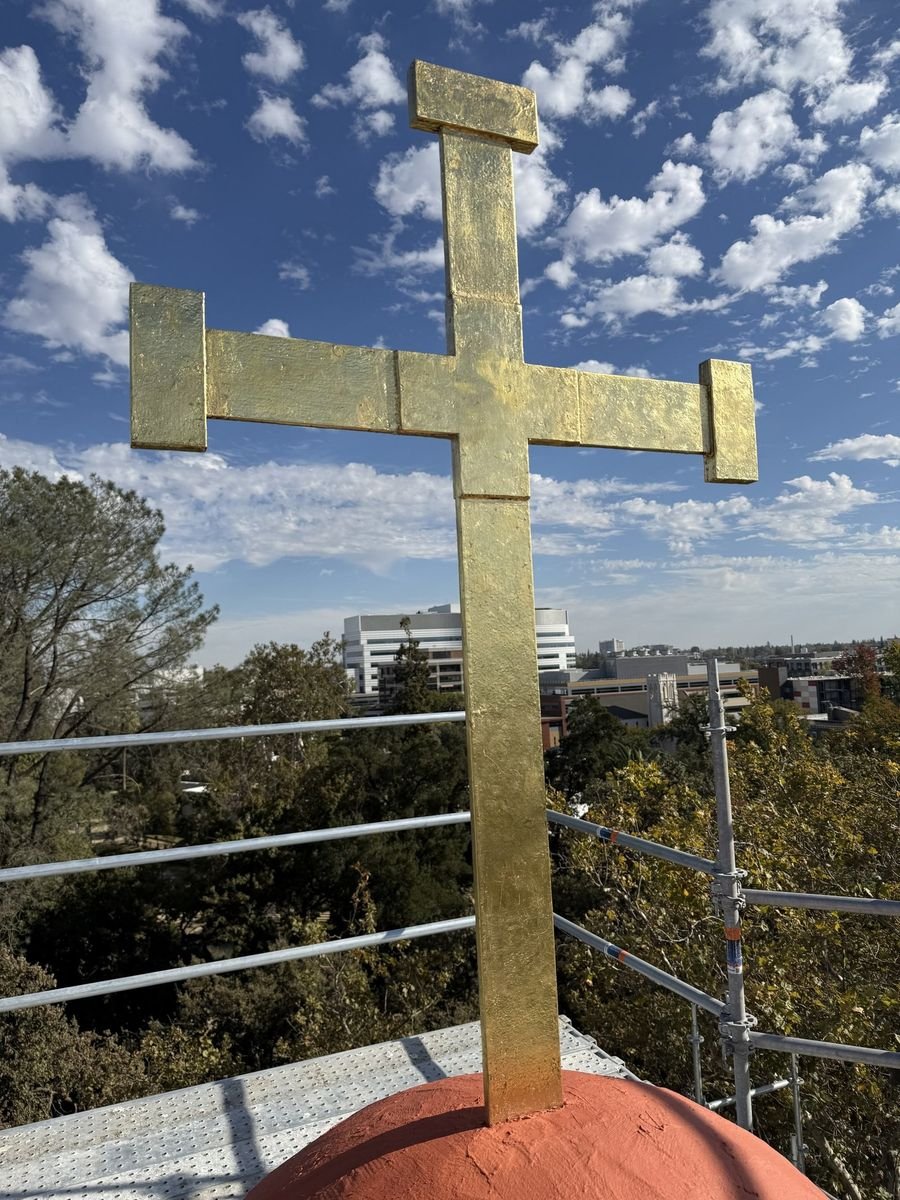 Gilded rooftop cross with squared arm ends against a blue sky and scattered clouds