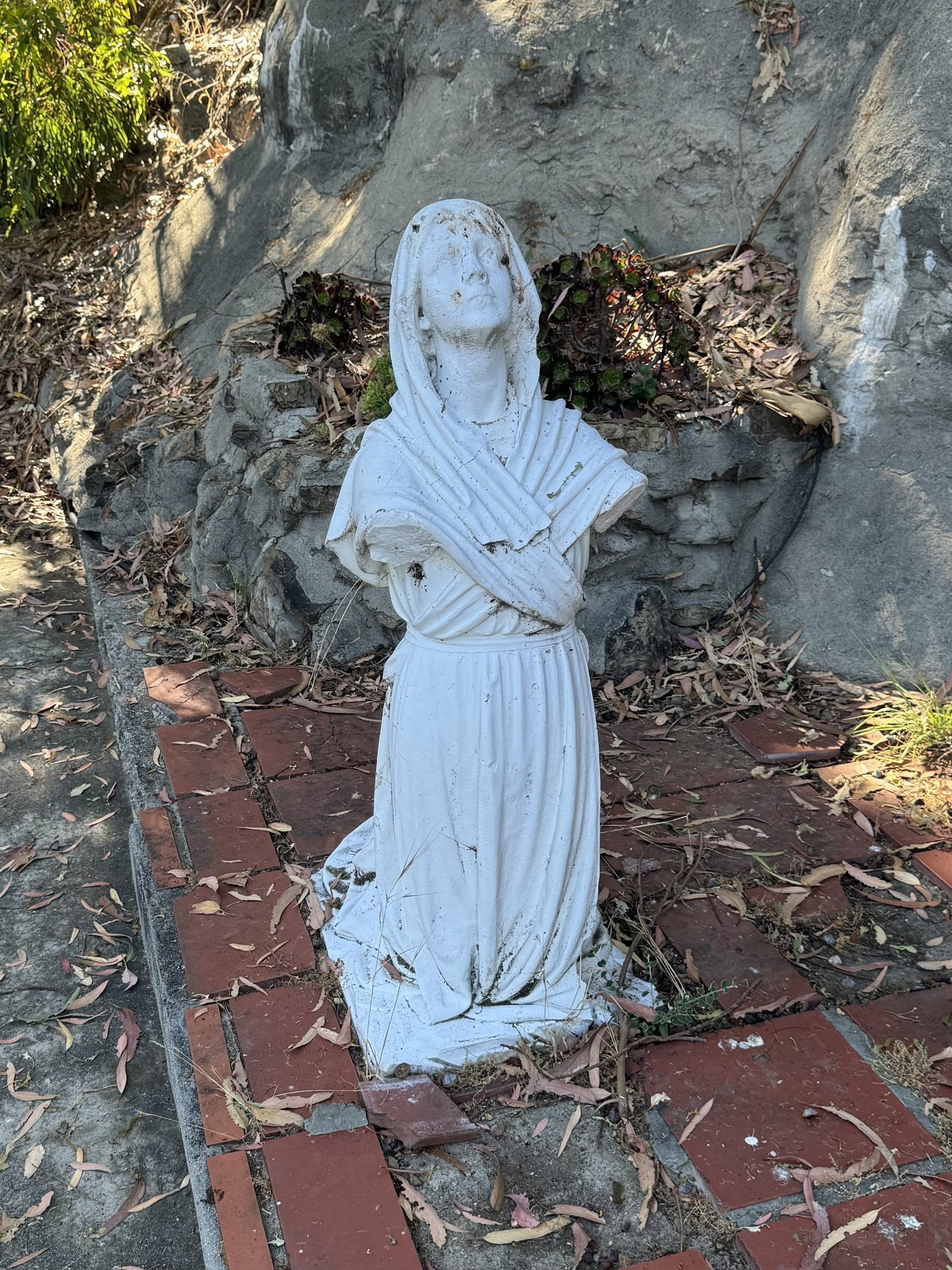Kneeling marble figure with missing forearms and hands in front of the grotto wall.