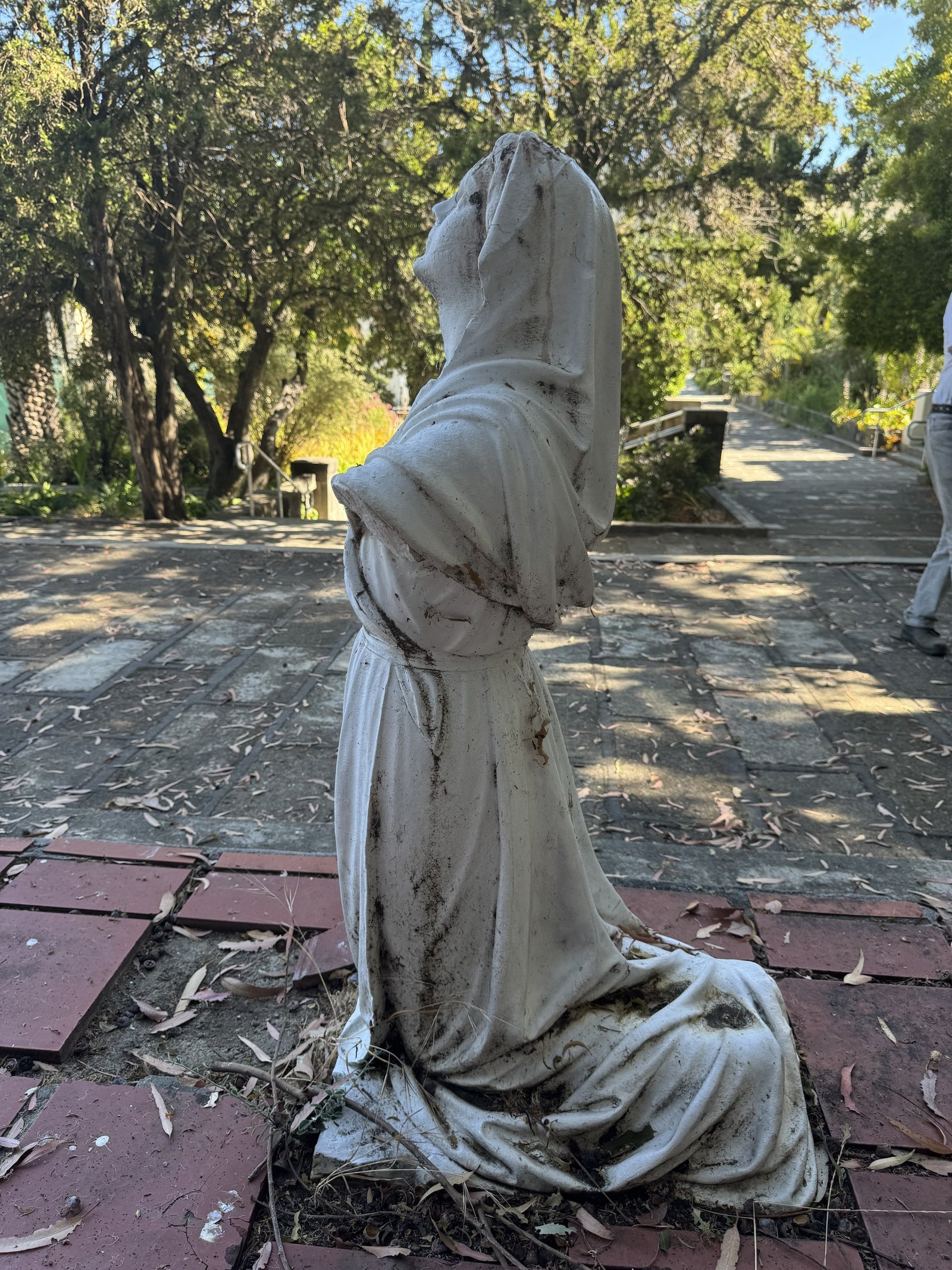 Rear three-quarter view of a weathered kneeling hooded marble figure on brick paving.