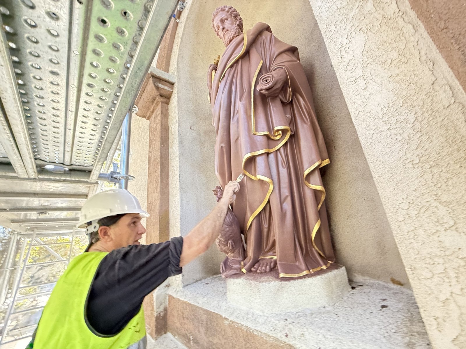Craftsperson on scaffolding working with a brush along gilded trim on a statue.