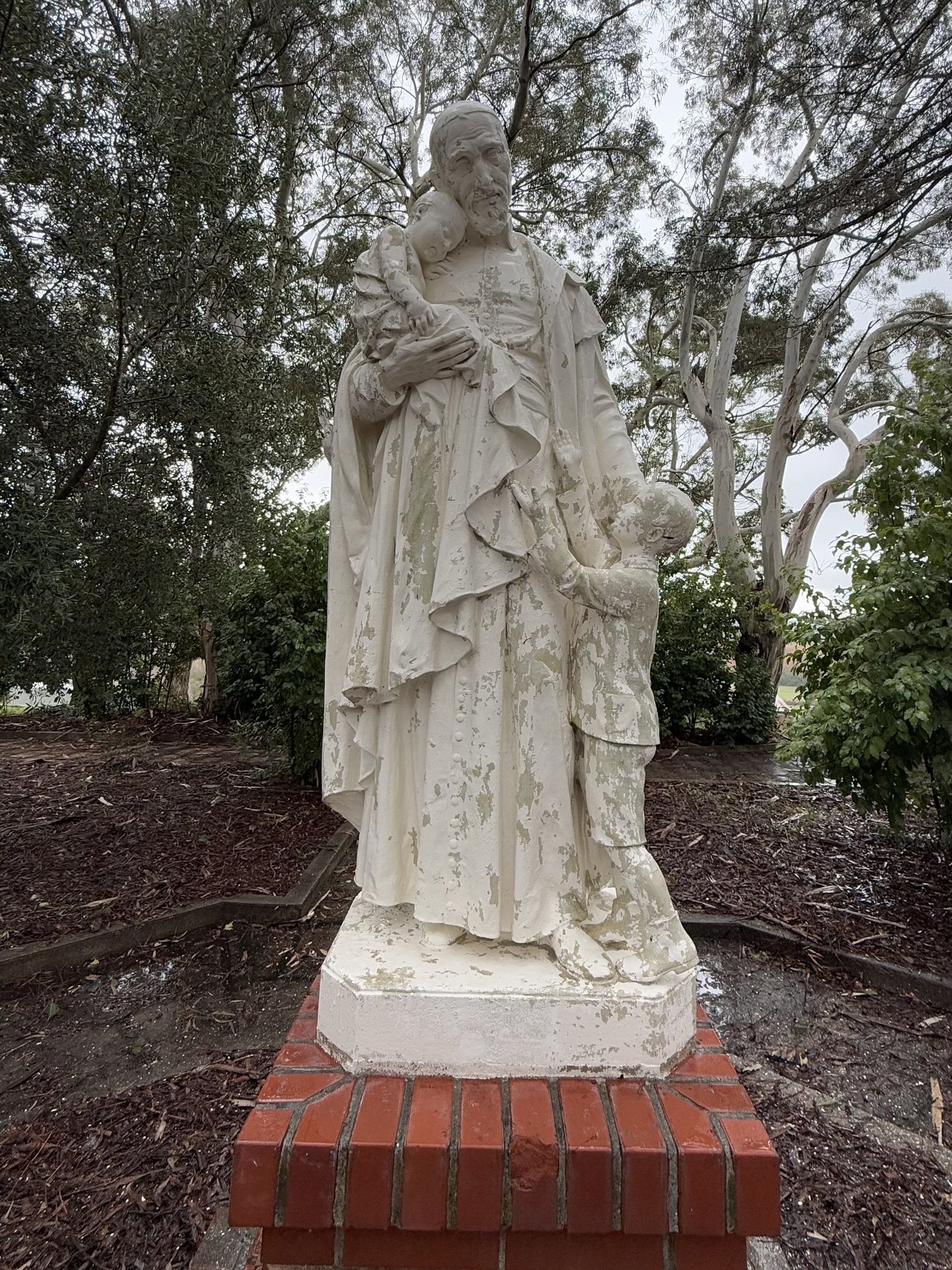 Standing sculpture holding a child with a second child beside him, with flaking finish across the robes.