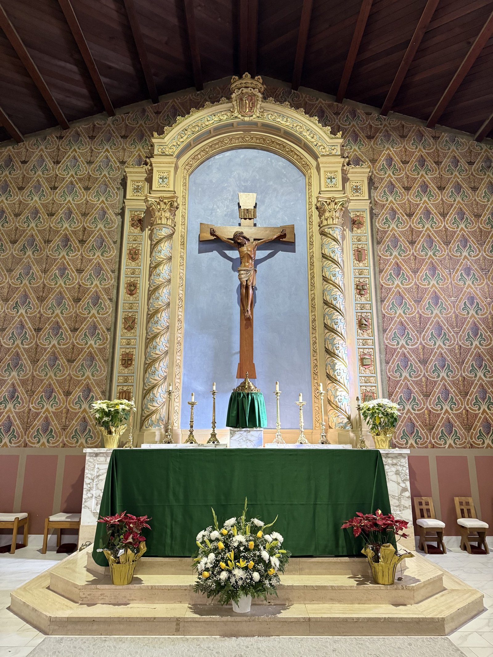Blue reredos niche and crucifix above an altar dressed in green, with flowers below.