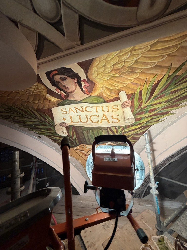 Repaired face of the winged figure above the SANCTUS LUCAS scroll, with a work light in front.