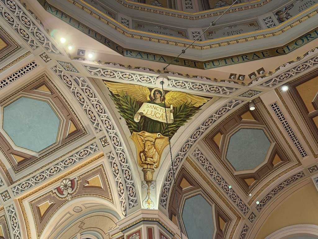 Triangular mural with a winged figure and scroll within the decorated cathedral ceiling.