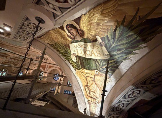 Angled view of the SANCTUS LUCAS mural with scaffold rails in front.