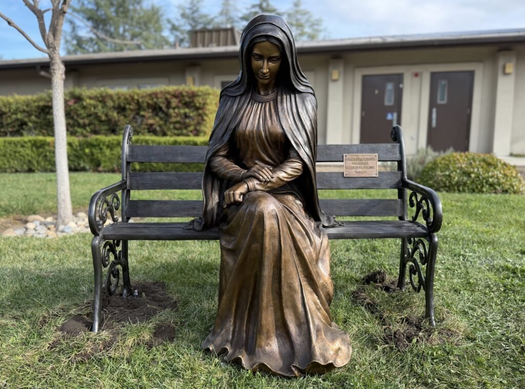 Full view of the bronze statue of Mary seated at one end of a bench with an open seat beside her.