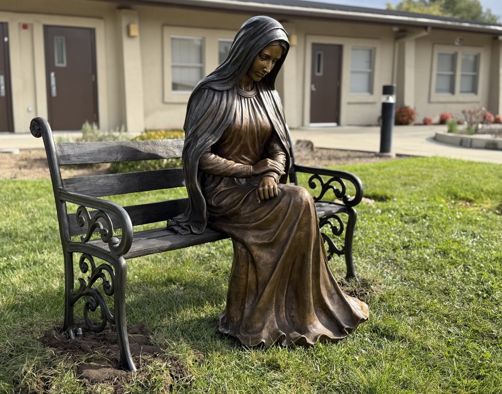 Side view of the bronze statue of Mary seated at one end of a slatted bench on grass.
