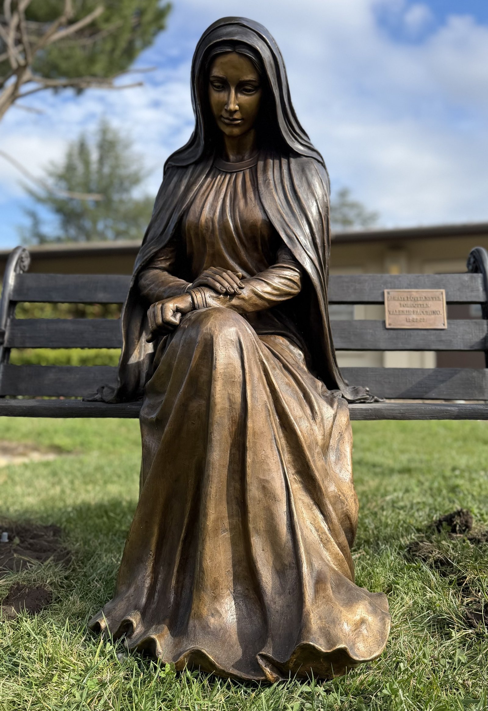 Front view of the bronze statue of Mary seated on an outdoor bench.