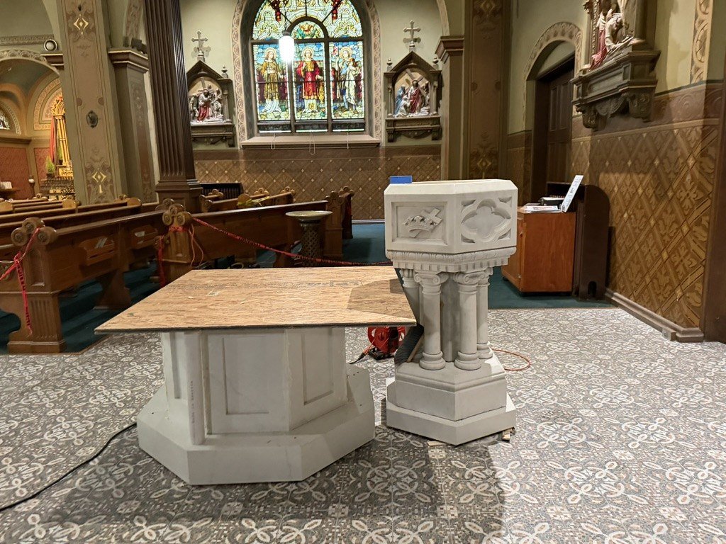 New octagonal basin base with a temporary plywood top beside the historic pedestal font on patterned tile.
